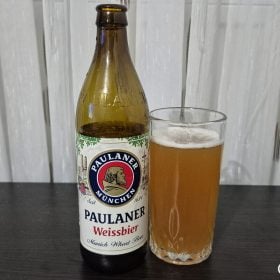 Chai bia Paulaner Weissbier cạnh ly bia vàng đục trên bàn gỗ