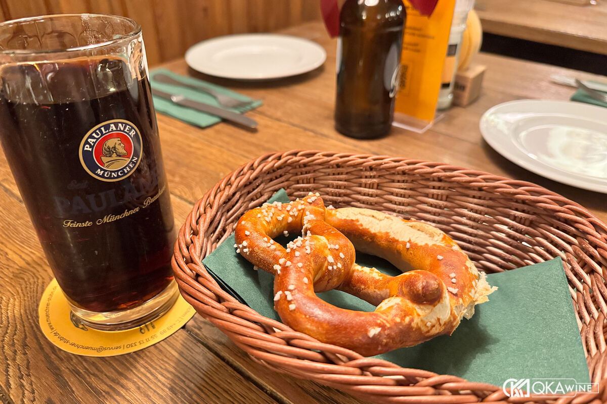 Ly bia Paulaner sẫm màu cạnh giỏ bánh pretzel trên bàn gỗ